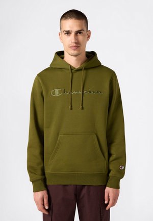 Man in een olijfgroene Champion-hoodie met een voorzak en borduursel, staande tegen een effen lichte achtergrond.