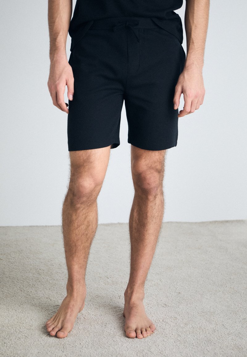 Shorts negre, până la mijlocul coapsei, cu un cordon la talie. Fabricate dintr-un material moale, cu o textură netedă și fără modele sau accente vizibile.