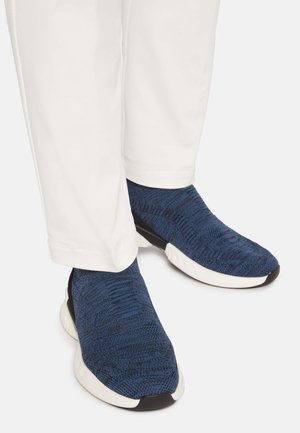 Blå strikkede sneakers med et tekstureret design, hvid sål og sorte detaljer, parret med hvide bukser.