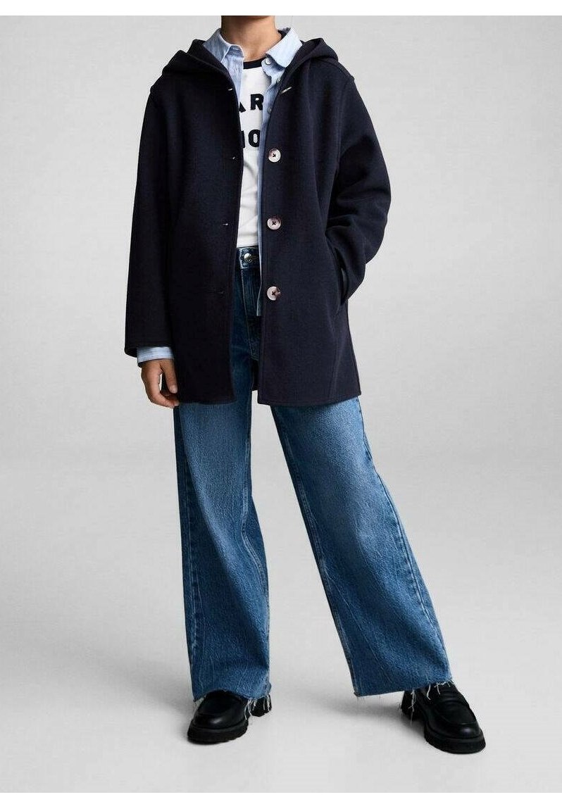 Námořnický kabát s kapucí a předním zapínáním na knoflíky, nošený přes bílou košili a modré džínové široké kalhoty, doplněný černými botami.