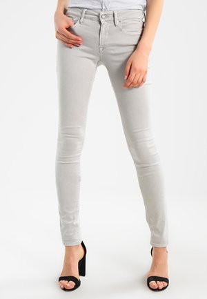 Kobieta ubrana w jasnoszare obcisłe jeansy i czarne sandały na wysokim obcasie, stojąca z jedną ręką w kieszeni, a drugą opartą na udzie.