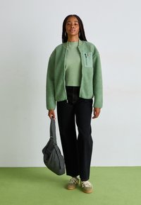 Green fleece jacket with a zip pocket, paired with a light green top and black wide-leg pants. Holds a checkered bag; wears beige sneakers.
