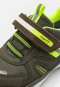 Zöld és szürke sportcipő, texturált szövetfelsővel, fényvisszaverő díszítéssel, tépőzárral és neon zöld részletekkel.