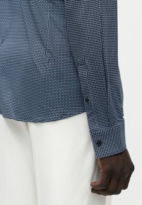 Κοντινό πλάνο ενός σκουρόχρωμου χεριού που φοράει μπλε μπλούζα με μικρά λευκά κυκλικά σχέδια και λευκό παντελόνι.