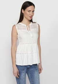 Blusa senza maniche bianca con dettagli in pizzo sulle spalle, chiusura a nodo al collo e texture a righe orizzontali. Vestibilità svasata con design a strati.