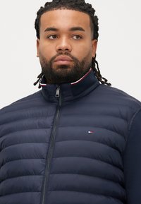 Mies, jolla on punotut hiukset ja joka on pukeutunut tummansiniseen tikattuun takkiin, jossa on pieni punainen, valkoinen ja sininen logo rinnassa, katsoo sivulle.