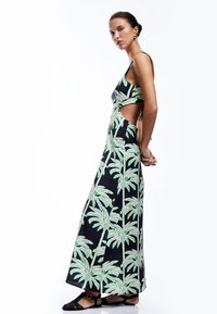 Schwarzes Maxikleid mit grünem Palmbaum-Muster, das über ausgeschnittene Seiten und dünne Träger verfügt. Texturierten Stoff mit fließendem Design.
