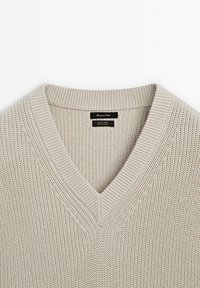 Pull en coton beige tricoté avec col en V et bordure côtelée, avec une étiquette noire à l'intérieur de l'encolure.