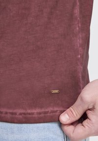 Burgunderrotes Baumwoll-T-Shirt mit einer glatten Textur, seitlichen Schlitzen und einem kleinen Metallschild mit der Aufschrift "ISOL.D" nahe dem Saum.