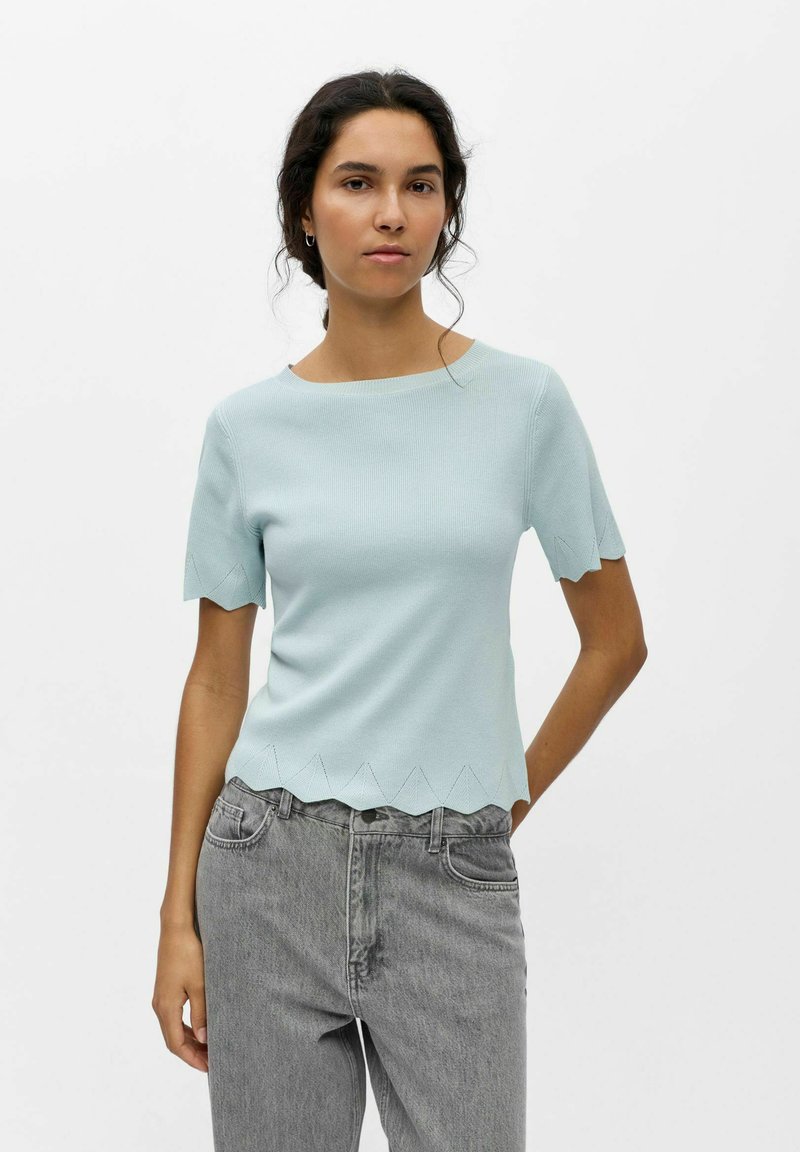 Femme aux cheveux foncés portant un haut bleu clair à manches courtes avec des bords festonnés et un jean gris taille haute, debout devant un fond uni.