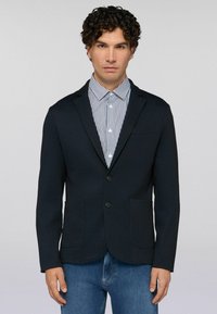 Muž s kudrnatými tmavými vlasy, oblečen�ý v tmavě modrém saku, pruhované košili s knoflíky a modrých džínách, na prostém světlém pozadí.