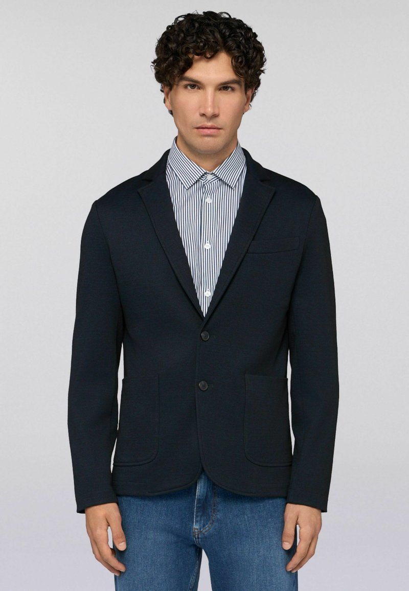 Muž s kudrnatými tmavými vlasy, oblečený v tmavě modrém saku, pruhované košili s knoflíky a modrých džínách, na prostém světlém pozadí.