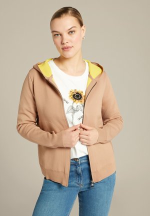 Bruine rits-hoodie met een gele binnenvoering, voorzien van een zachte textuur en zijzakken, gedragen over een wit shirt met een zonnebloemontwerp.