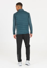 Veste de sport turquoise avec dos rembourré, manches longues et col montant, assortie à un pantalon noir fuselé et des sneakers gris clair.
