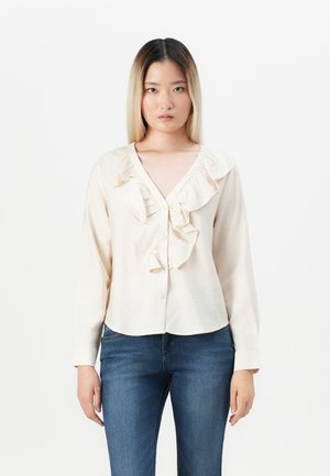 Kremfarget skjorte med knappestolpe og volanger foran, lange ermer og myk tekstur, kombinert med mørkeblå jeans.