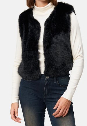 Femme portant une veste noire en fausse fourrure par-dessus un col roulé blanc et un jean bleu foncé, debout devant un fond clair uni.