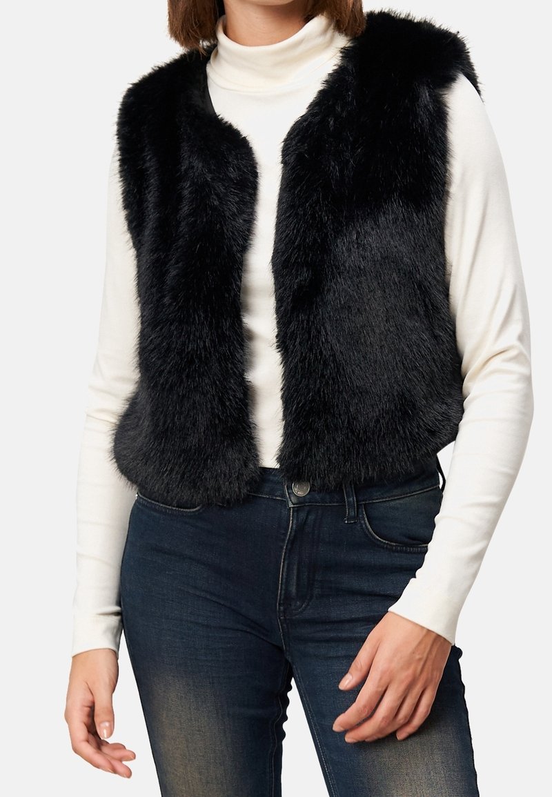 Femme portant une veste noire en fausse fourrure par-dessus un col roulé blanc et un jean bleu foncé, debout devant un fond clair uni.
