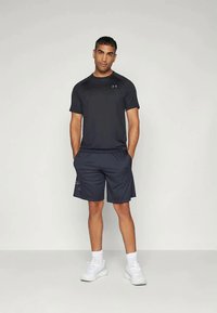 Uomo in piedi con le mani in tasca, indossa una maglietta nera Under Armour, pantaloni corti neri, calzini bianchi e sneakers bianche su uno sfondo neutro.