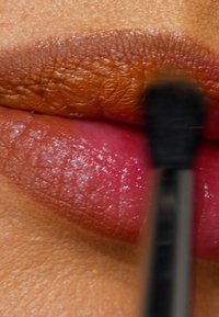 Labbra lucide in due tonalità: labbro superiore marrone opaco e labbro inferiore rosa lucido, con un piccolo pennello che applica il prodotto sulla zona marrone.