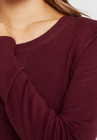 Maglione bordeaux con scollatura e polsini a coste, caratterizzato da una consistenza morbida e una leggera elasticità per il massimo comfort.