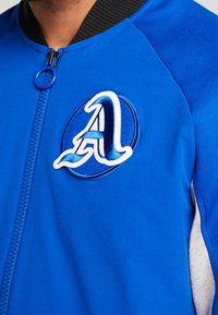 Sudadera azul con un cuello de canalé negro, que presenta un logo bordado circular en azul y blanco. Cierre de cremallera en el frontal.