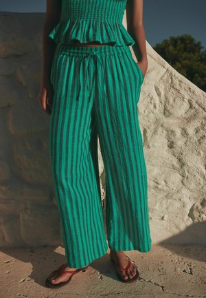 Pantaloni largi cu dungi verticale verzi și negre, cu șnur, asortați cu un top ruched asortat, purtați cu șlapi roșii pe fundal de zid de piatră.