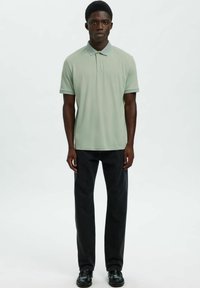 Svetlo zelena polo majica s kratkimi rokavi, ravnim pletenim ovratnikom in stranskimi razporki, kombinirana s črnimi kavbojkami z ravnimi hlačnicami in črnimi čevlji.