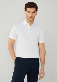 Polo bianca con colletto, maniche corte e un logo ricamato discreto sul petto, abbinata a pantaloni scuri.