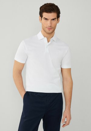 Vit polo med krage, korta ärmar och en subtil broderad logotyp på bröstet, i kombination med mörka byxor.