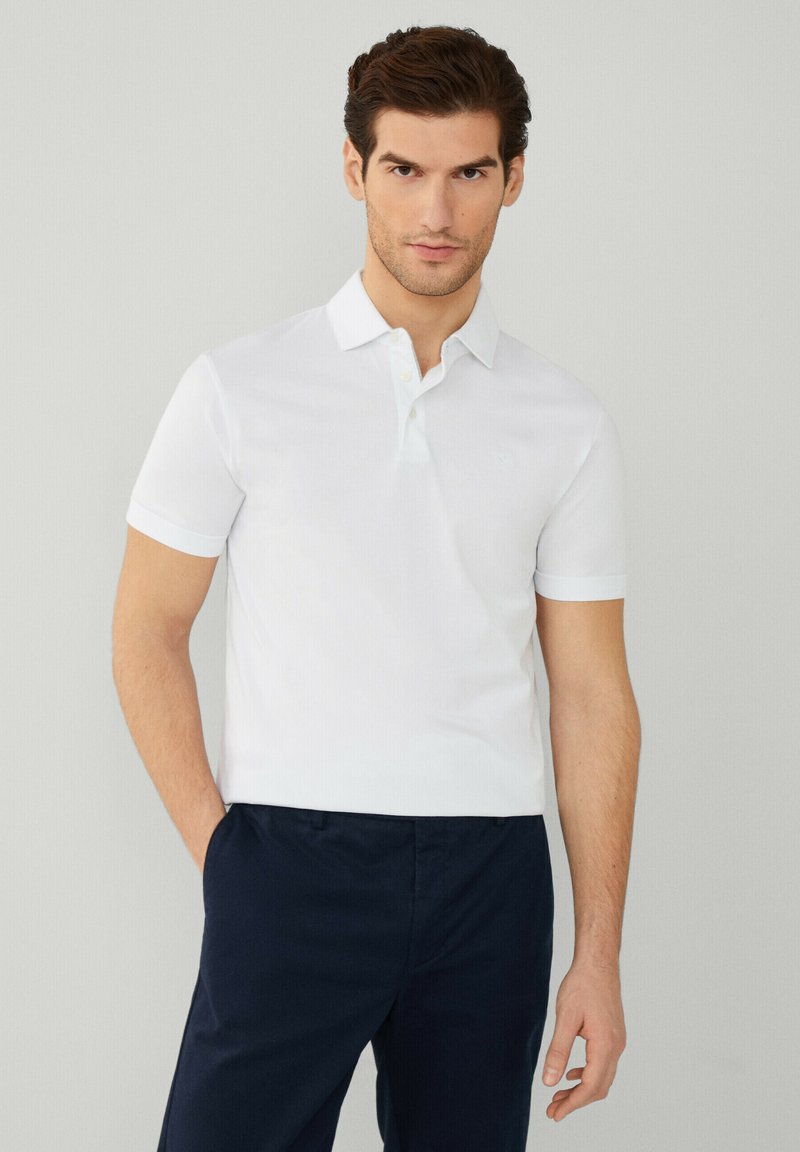 Polo bianca con colletto, maniche corte e un logo ricamato discreto sul petto, abbinata a pantaloni scuri.