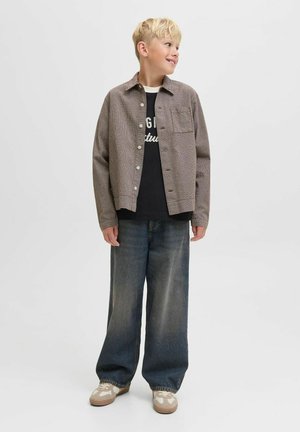 Chemise à carreaux marron sur un t-shirt graphique noir, associée à un jean en denim foncé à jambes larges et des baskets beiges avec des accents blancs.