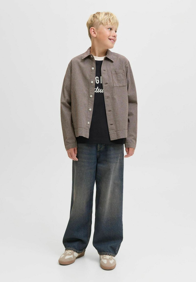 Rutigt brunt skjorta över en svart t-shirt med tryck, kombinerat med vida mörk denimjeans och beige sneakers med vita detaljer.