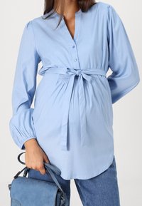 Svetle modrá košile s dlouhým rukávem, s knoflíkovým límečkem a stahovací šňůrkou v pase. Hladká látka s nabíranými rukávy a volným střihem.