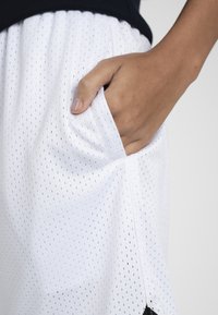 Main reposant dans la poche latérale d'un short de sport en mesh blanc avec ceinture élastique sur fond uni.