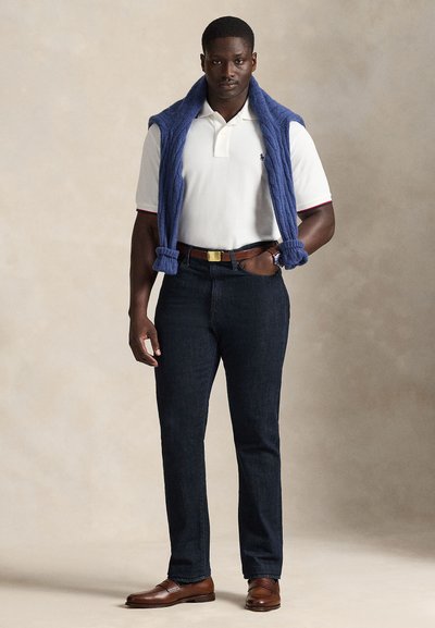 Bărbat purtând un tricou polo alb, blugi închiși la culoare, curea și pantofi maro, cu un pulover albastru pe umeri, stând pe un fundal neutru.