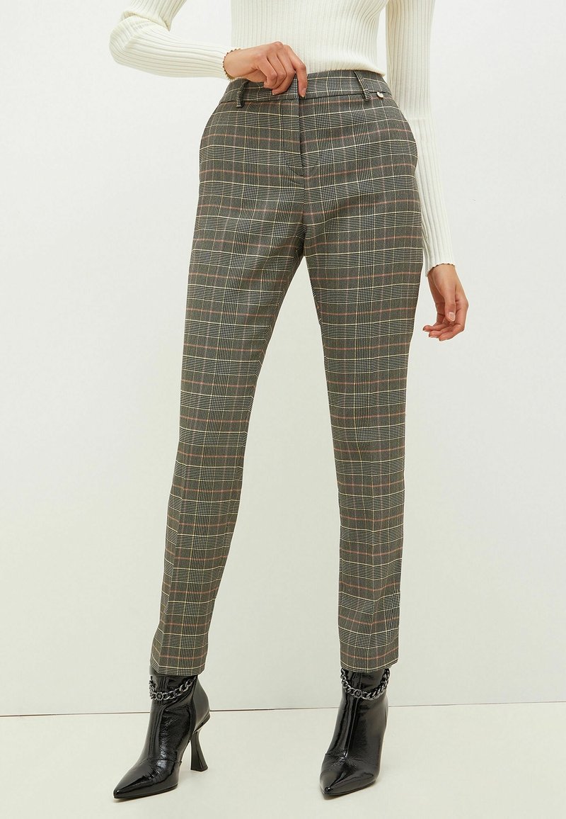 Pantalon à carreaux gris avec une coupe ajustée, une taille mi-haute, et des lignes subtiles rose et blanche ; associé à des bottines noires à bouts pointus.