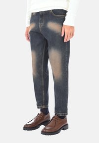 Jeans blu scuro con macchie scolorite, taglio dritto e design standard a cinque tasche, abbinati a scarpe di pelle marroni e calzini blu navy.