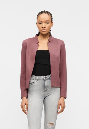 Vrouw met gevlochten haar, gekleed in een mauve cropped blazer, zwarte strapless top en lichtgrijze skinny jeans met scheuren, staand tegen een witte achtergrond.