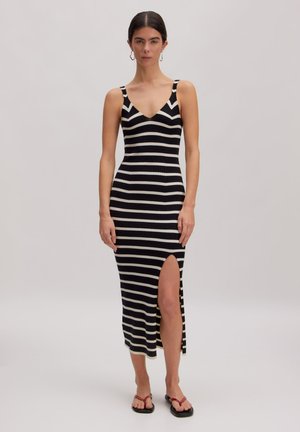 Femme portant une robe sans manches rayée noir et blanc horizontale avec une fente haute à la cuisse, debout devant un fond uni.