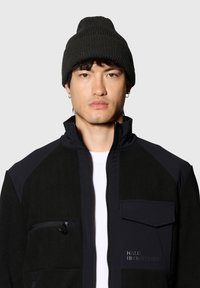 Schwarze Zip-up-Jacke aus weichem Material mit kontrastierenden Einsätzen, einer Reißverschlusstasche und einem kleinen Logo auf der Brust. Getragen mit einer schwarzen Mütze.