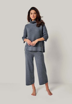 Frau steht barfuß und trägt einen lockeren grauen gerippten Rollkragenpullover und weite graue Hosen vor einem schlichten Hintergrund.