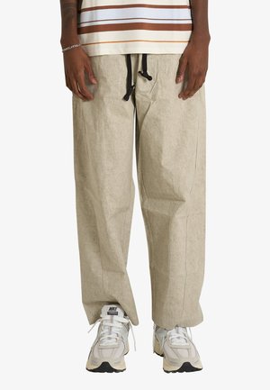 Persona che indossa pantaloni beige a gamba larga con cordini neri e sneakers bianche Nike, in piedi davanti a uno sfondo bianco uniforme.