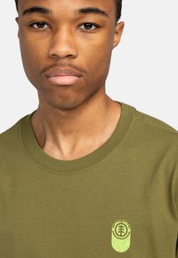 Camiseta verde oliva con cuello redondo y mangas cortas, que presenta un pequeño logotipo verde claro en la zona izquierda del pecho. Textura de tela suave.