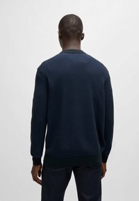 Homme aux cheveux courts portant un pull texturé bleu foncé et un pantalon foncé, vu de dos sur un fond clair uni.