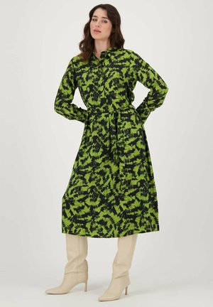 Robe à motifs verts et noirs, manches longues, col, taille ceinturée, longueur midi, associée à des bottes beiges montantes.