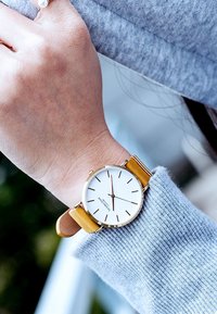 Reloj de pulsera en tono dorado con una esfera blanca, marcadores de hora negros y una correa de cuero amarilla, usado en una mano que lleva un suéter de punto gris.