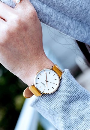 Guldtonet armbåndsur med en hvid urskive, sorte timemarkører og et gult læderrem, der bæres på en hånd i en grå striktrøje.