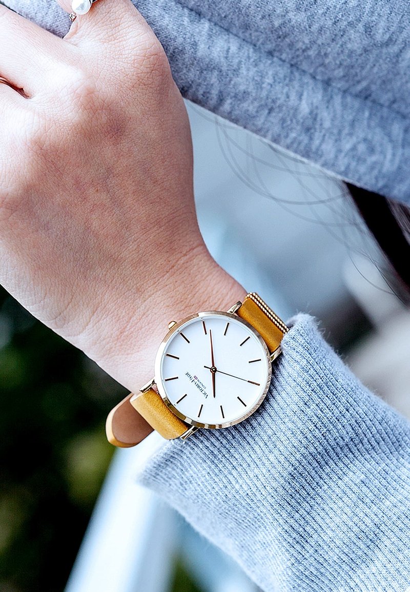 Reloj de pulsera en tono dorado con una esfera blanca, marcadores de hora negros y una correa de cuero amarilla, usado en una mano que lleva un suéter de punto gris.
