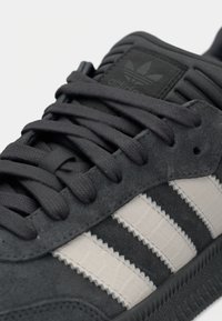 Γκρι σουέντ αθλητικό παπούτσι με υφή στη μαύρη σόλα, μαύρα κορδόνια και τρεις λευκές ανάγλυφες ρίγες στο πλάι. Σημαντική ετικέτα με το λογότυπο στη γλώσσα.