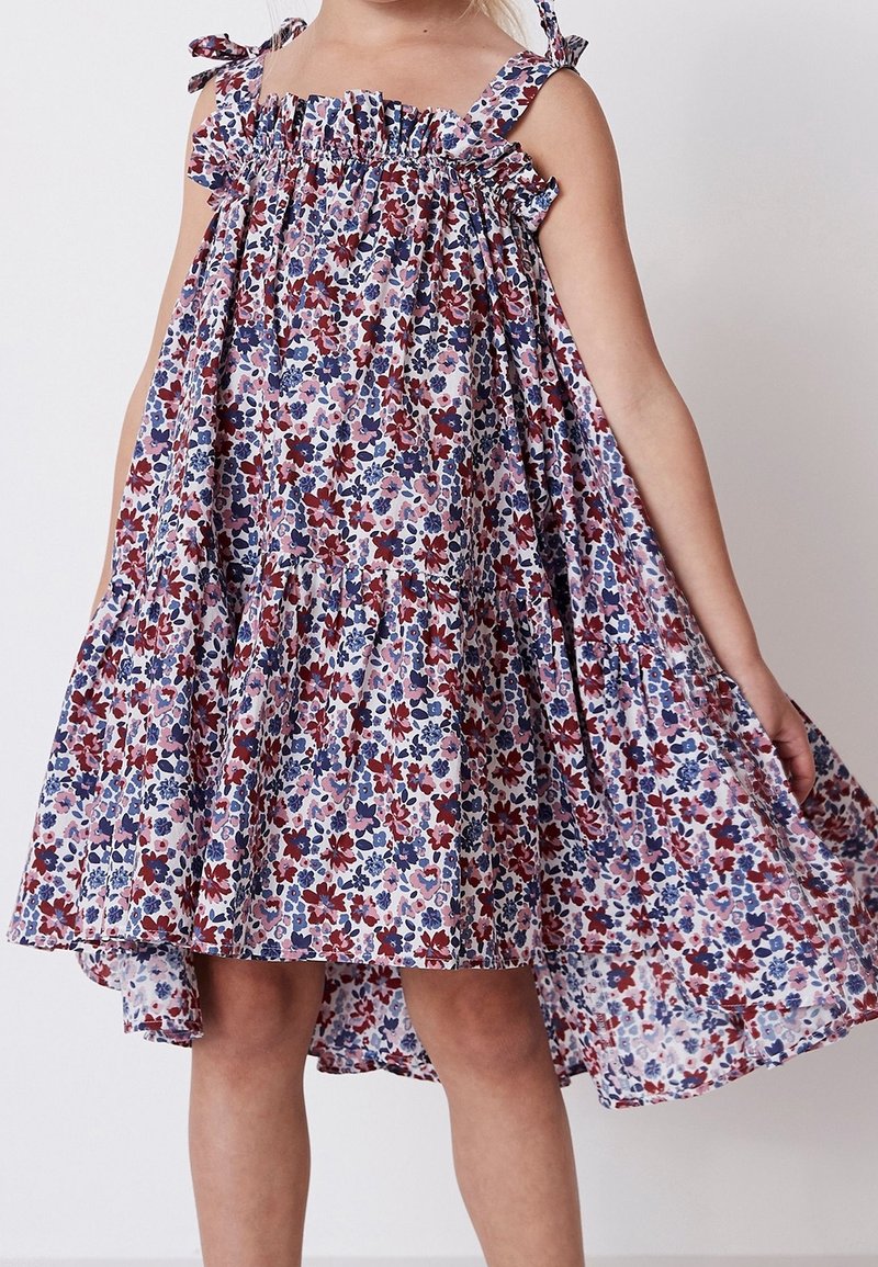 Jeune enfant portant une robe sans manches à imprimé floral avec des bretelles à volants et une jupe fluide sur un fond clair uni.